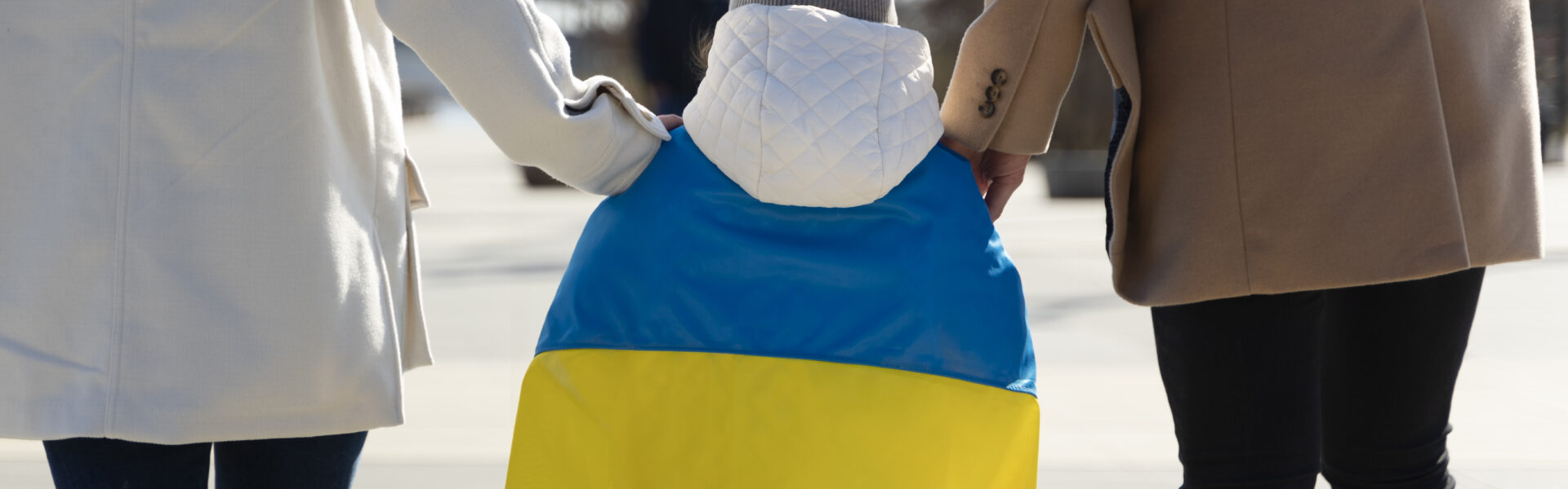 idąca rodzina fotografowana z tyłu. Pomiedzy rodzicami dziecko około 5-7 lat. Rodzice trzymaja dziecko za ręce. Dziecko na plecach okryte jest flagą Ukrainy
