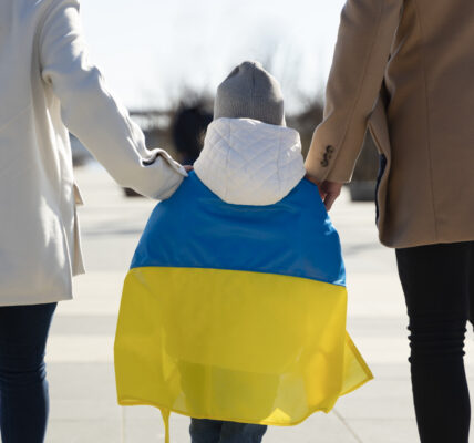 idąca rodzina fotografowana z tyłu. Pomiedzy rodzicami dziecko około 5-7 lat. Rodzice trzymaja dziecko za ręce. Dziecko na plecach okryte jest flagą Ukrainy